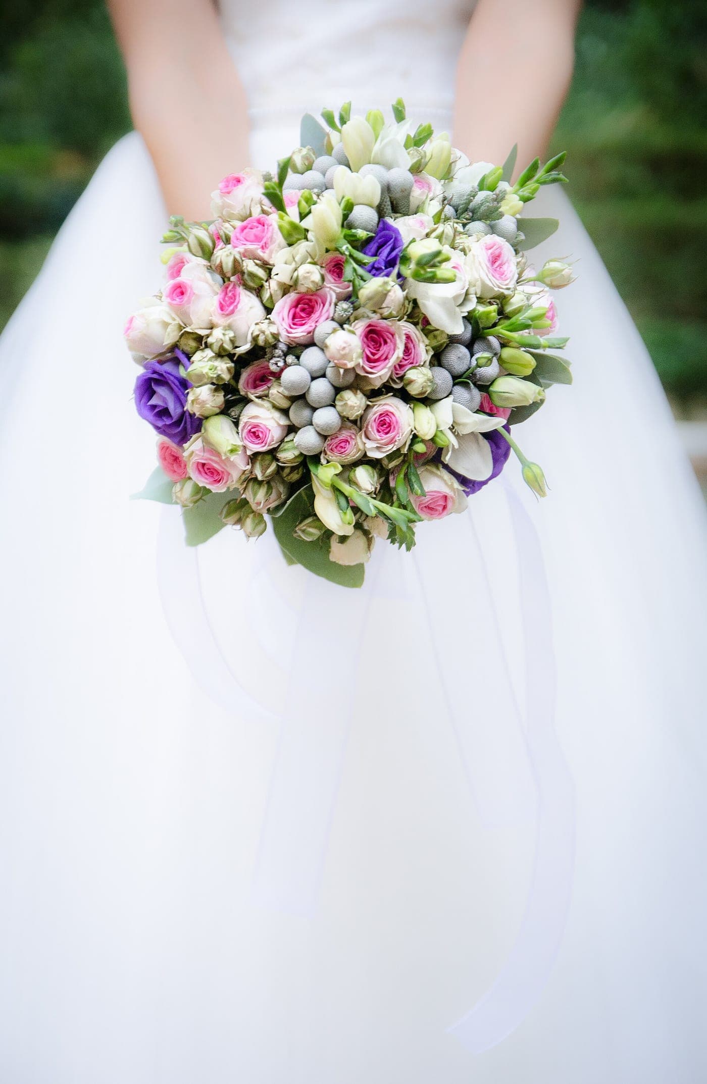A round bouquet of pink spray roses, silver brunia and purple lisianthus held against a tulle gown.
