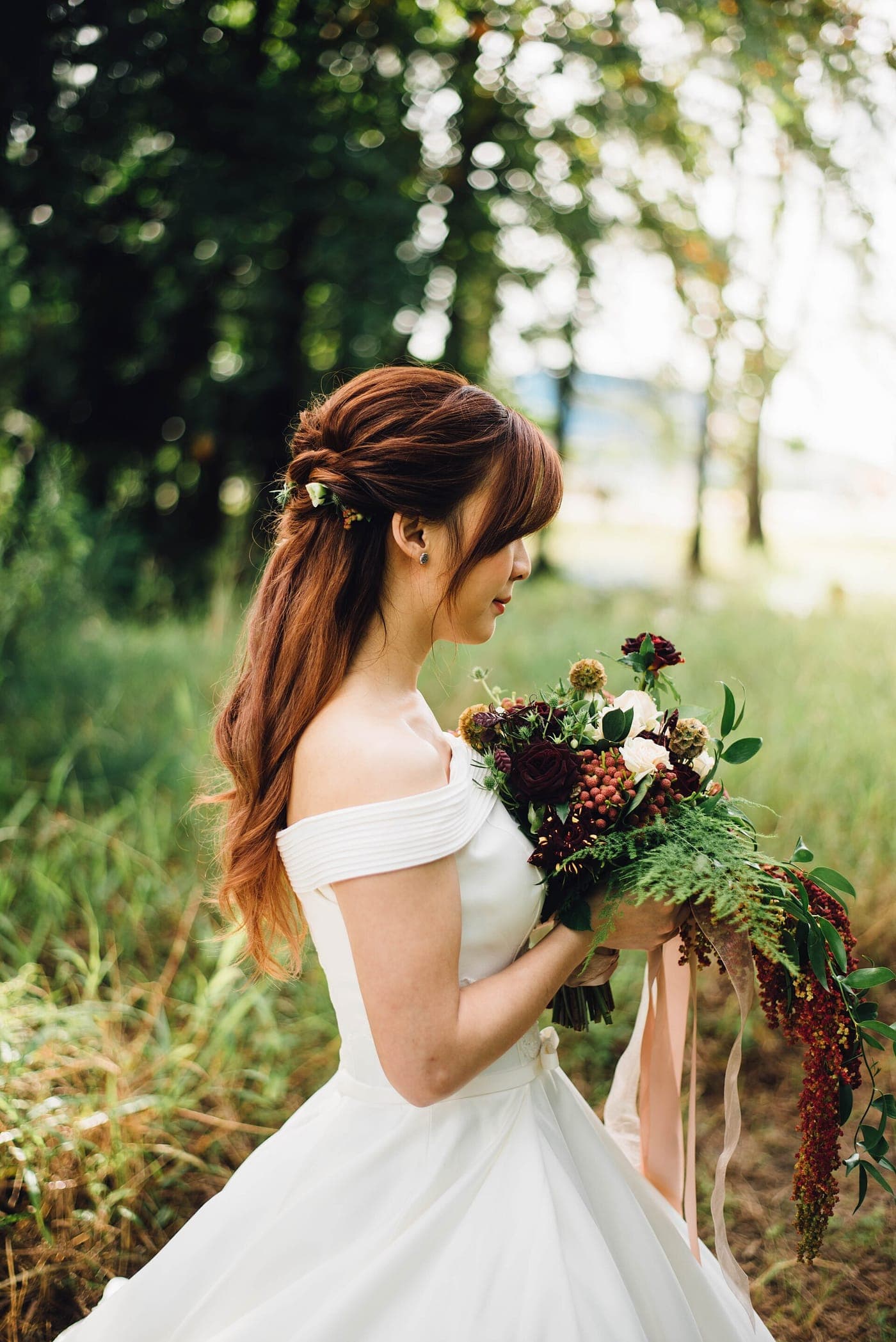 A bride in an off-the-shoulder gown holding a loose, wild bouquet in a summer meadow.