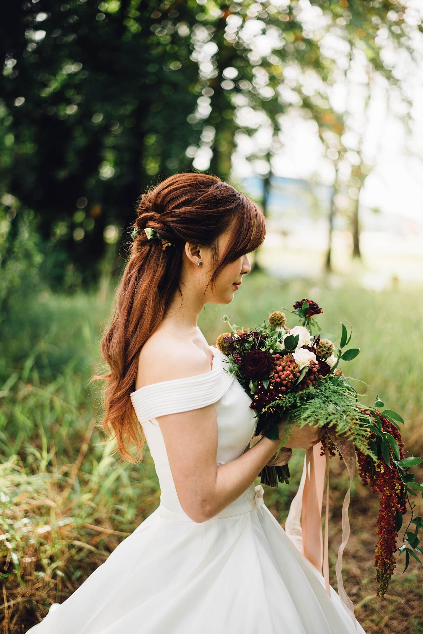 A bride in an off-the-shoulder gown holding a loose, wild bouquet in a summer meadow.