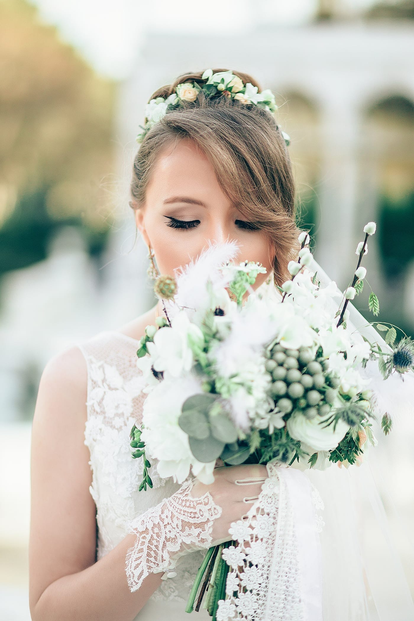 A bride with a small flower crown looking down at a white and green bouquet with eucalyptus and pussy willow.
