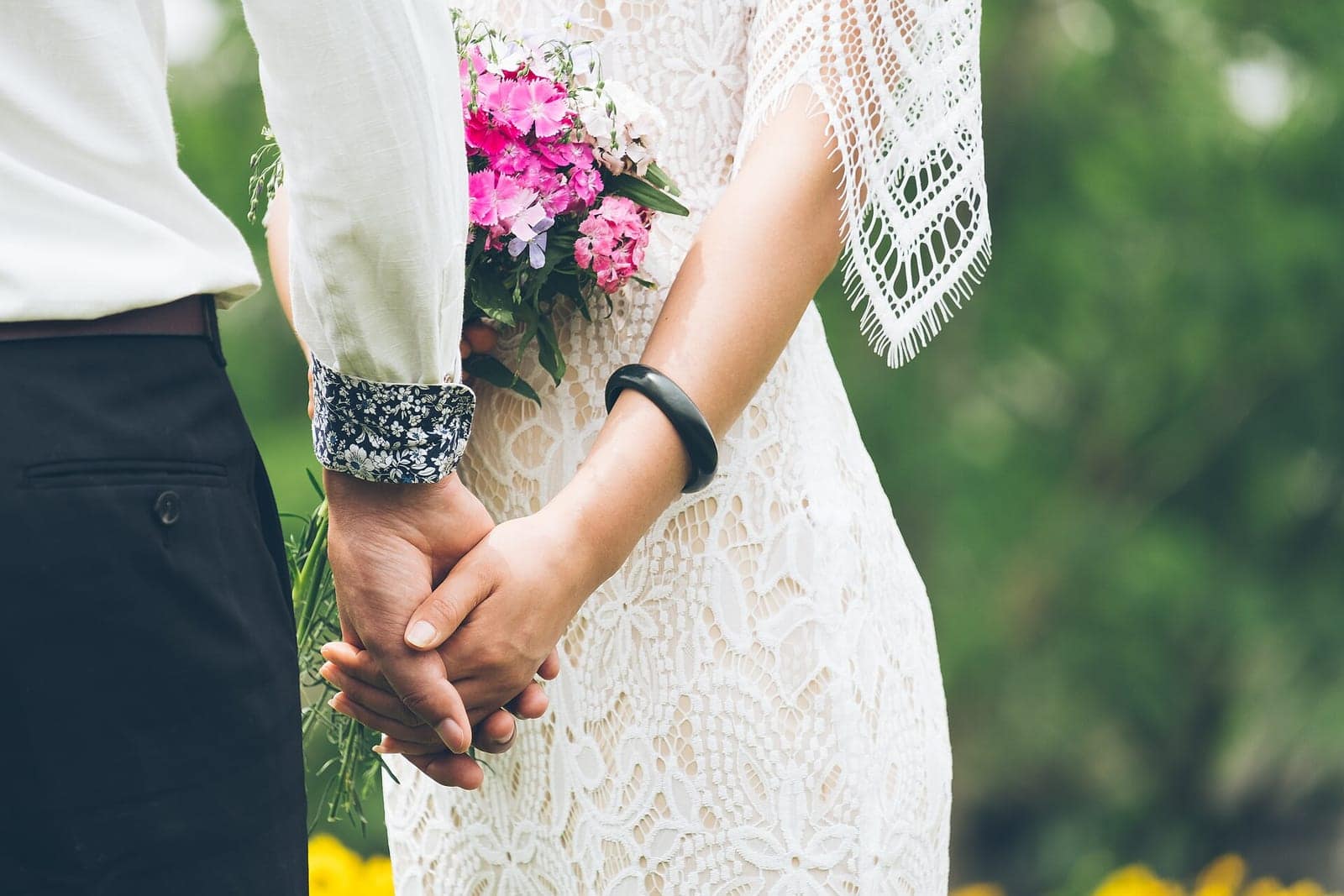 A couple holding hands, the bride in a lace dress carrying a small pink posy.