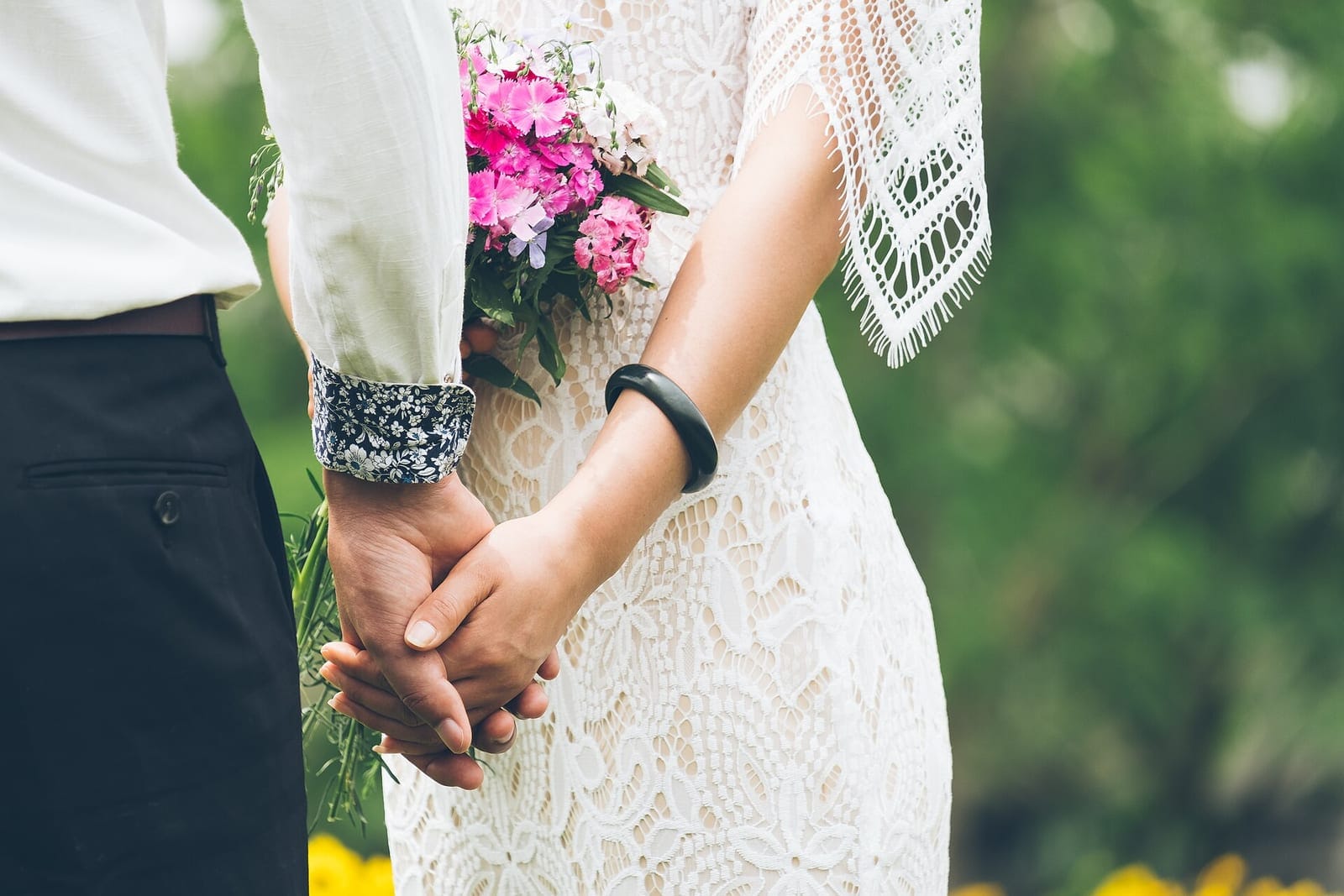 A couple holding hands, the bride in a lace dress carrying a small pink posy.