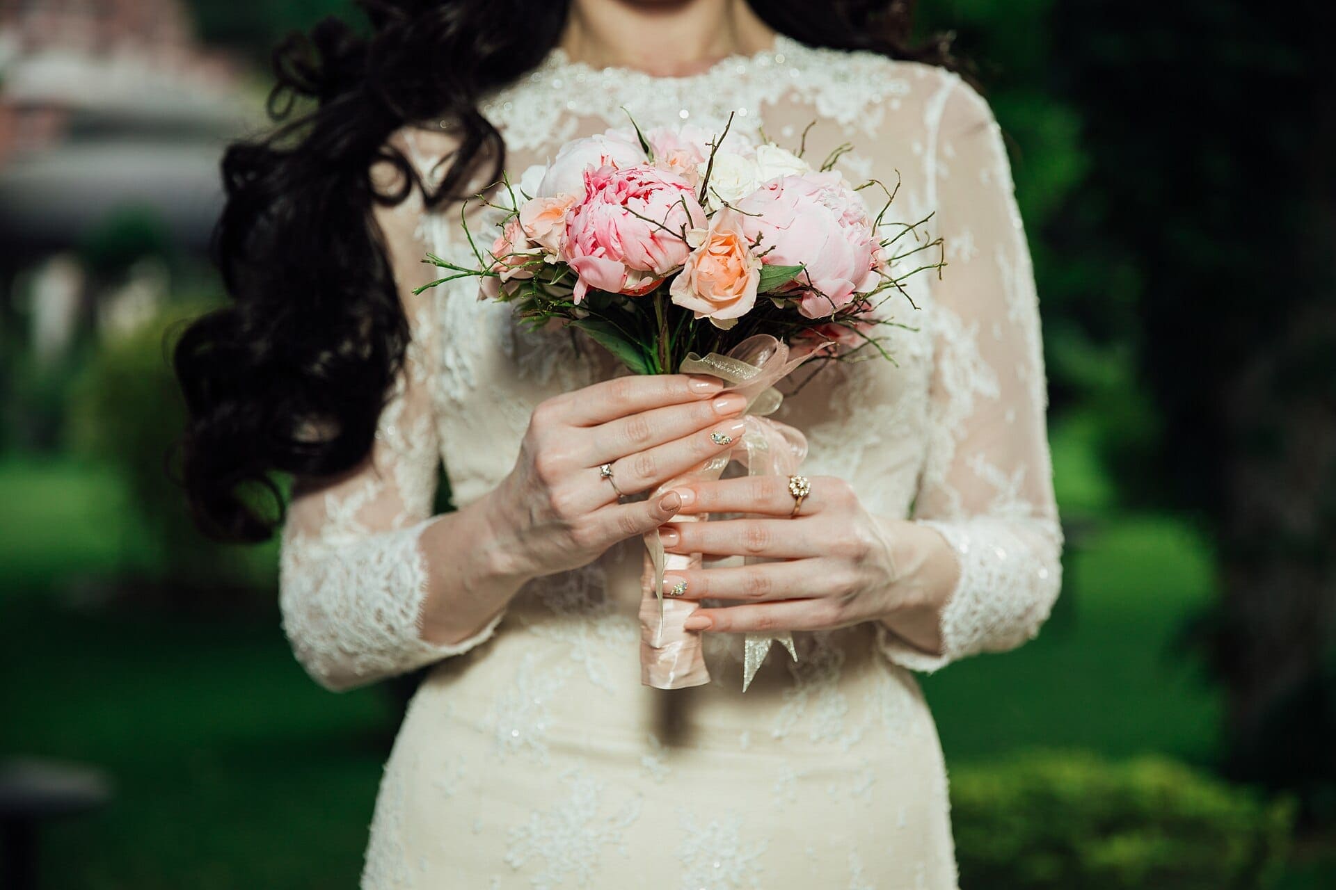 A bride in a lace gown holding a small bouquet of blush garden roses against deep green foliage.